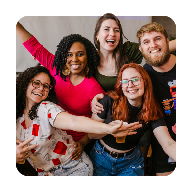 Grupo de pessoas com características diversas abrindo os braços e sorrindo. À esquerda temos uma mulher branca de cabelos cacheados e óculos de grau, com uma roupa de estampa e calça jeans. Ao lado dela temos uma mulher de pele negra, vestindo uma blusa rosa e um brinco de mandala. Um pouco abaixo temos outra mulher branca, com cabelos ruivos, vestindo uma blusa preta e calça jeans. Acima uma mulher de blusa verde musgo, branca, de cabelo médio e loiro, e ao lado dela um homem com barba e cabelos ruivos, com detalhes brancos. Sua camiseta é de personagem.