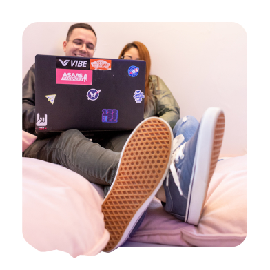 Homem e mulher sentados em poffs brancos mexendo no computador. O Homem está usando uma calça verde, tênis azul e está sorrindo. A mulher tem o cabelo ruivo, está usando uma blusa verde e seu rosto está tampado pela tela do computador.